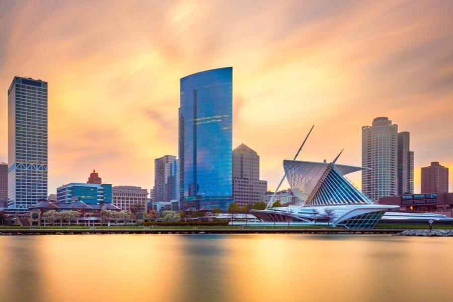 An image of tall city buildings reflecting off a calm lake in the foreground, with a clear sky above.